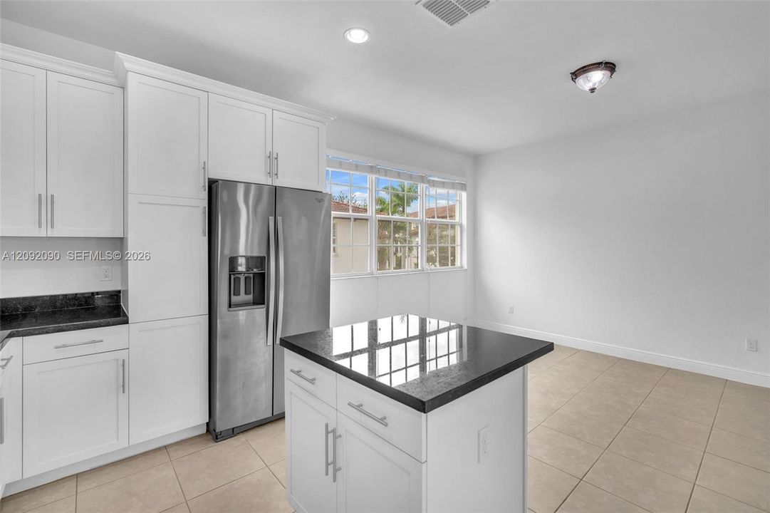 White shaker-style cabinetry, dark stone countertops, central kitchen island, stainless steel refrigerator, and ceramic tile flooring