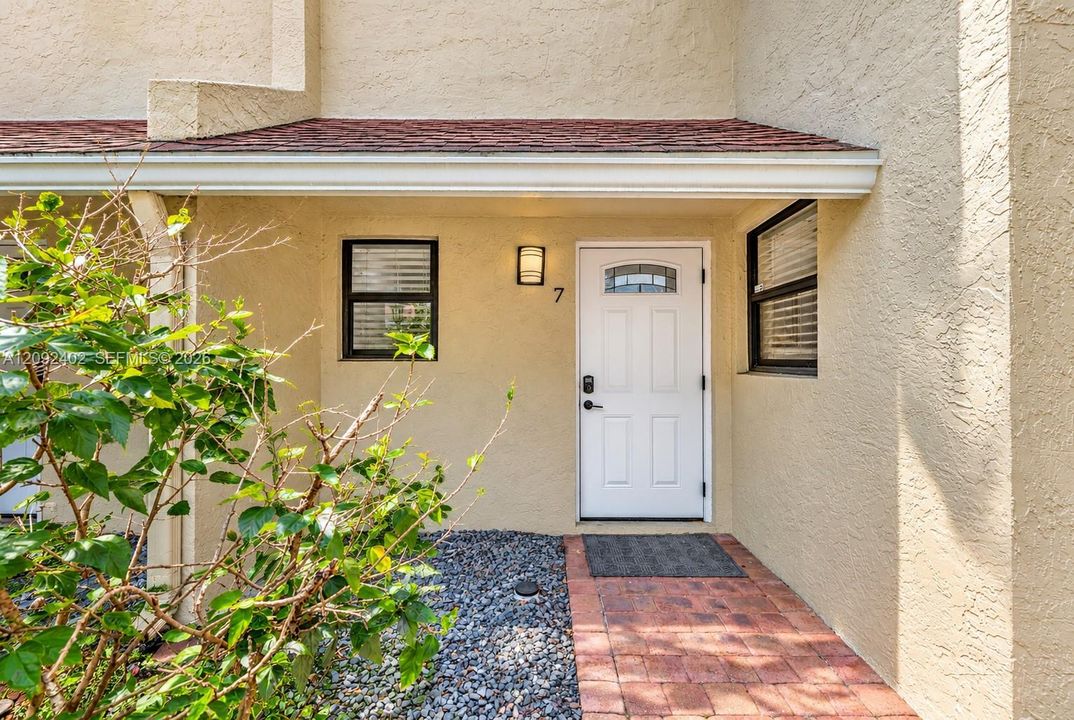Stucco exterior entrance featuring a brick-paver walkway, red shingle roof overhang, white door with a glass inlay, and wall-mounted exterior lighting