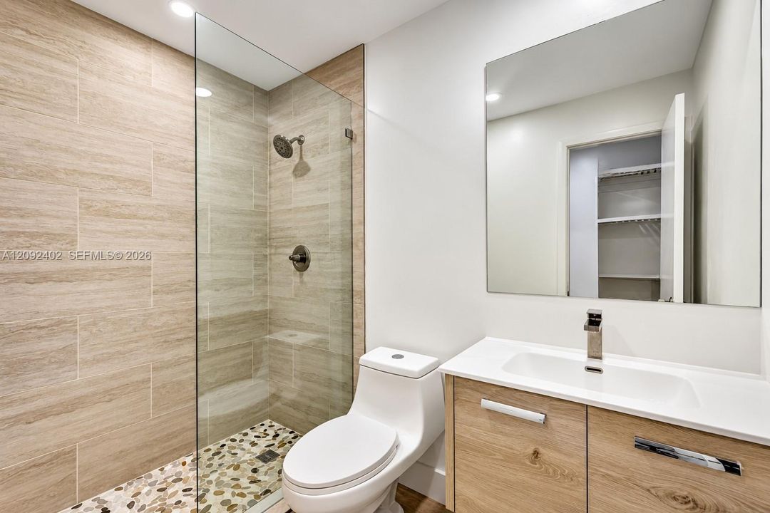 Bathroom featuring a glass-enclosed shower with pebble-stone flooring and large-format wall tile, wood-finish vanity, solid-surface countertop with integrated sink, and polished chrome fixtures