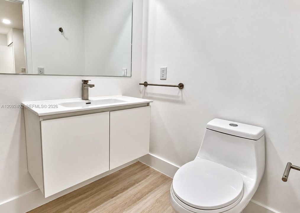 Floating vanity with a white countertop and integrated sink, wall-mounted faucet, expansive frameless mirror, one-piece dual-flush toilet, and wood-finish flooring