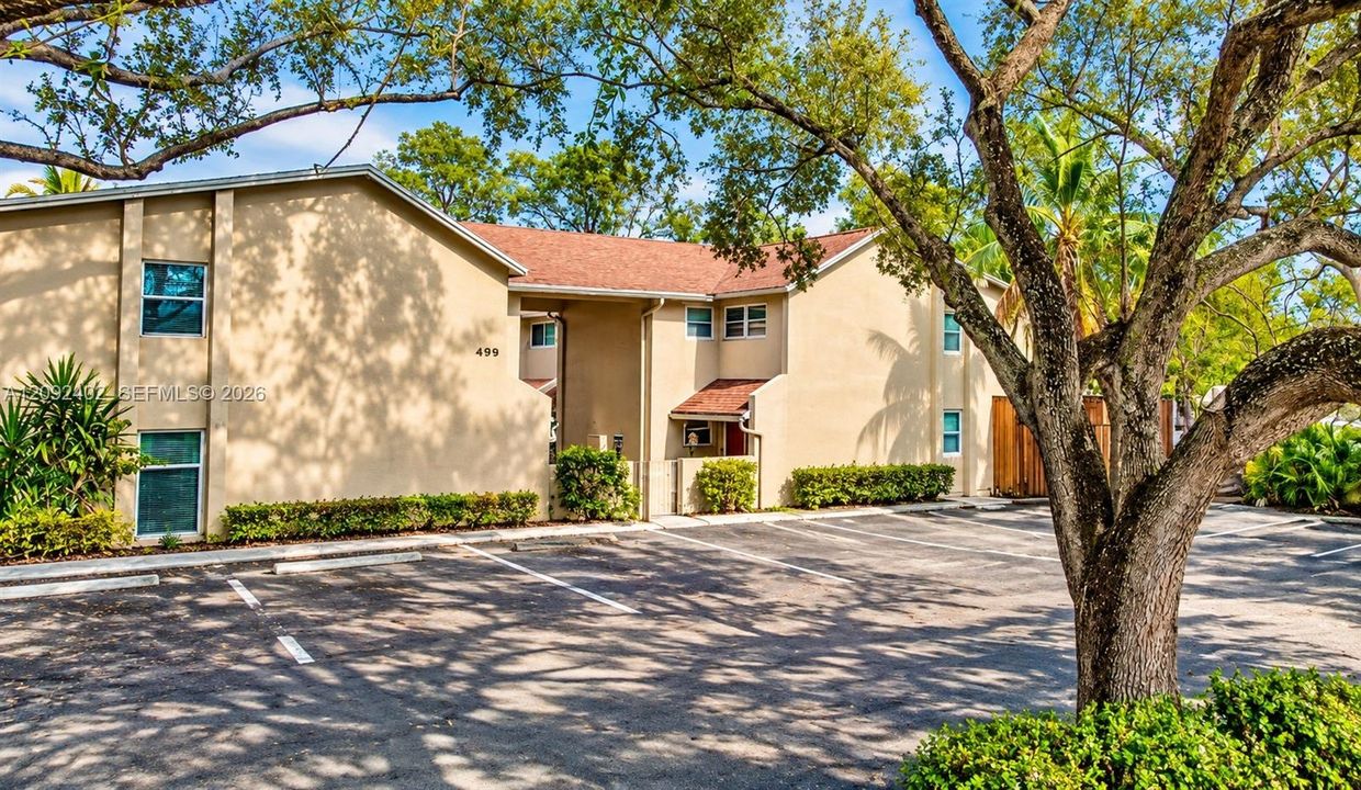 Some landscaping visually staged for illustrative purposes.  Exterior building facade featuring a tan stucco finish, multi-pitched shingled roof, asphalt-paved parking area.