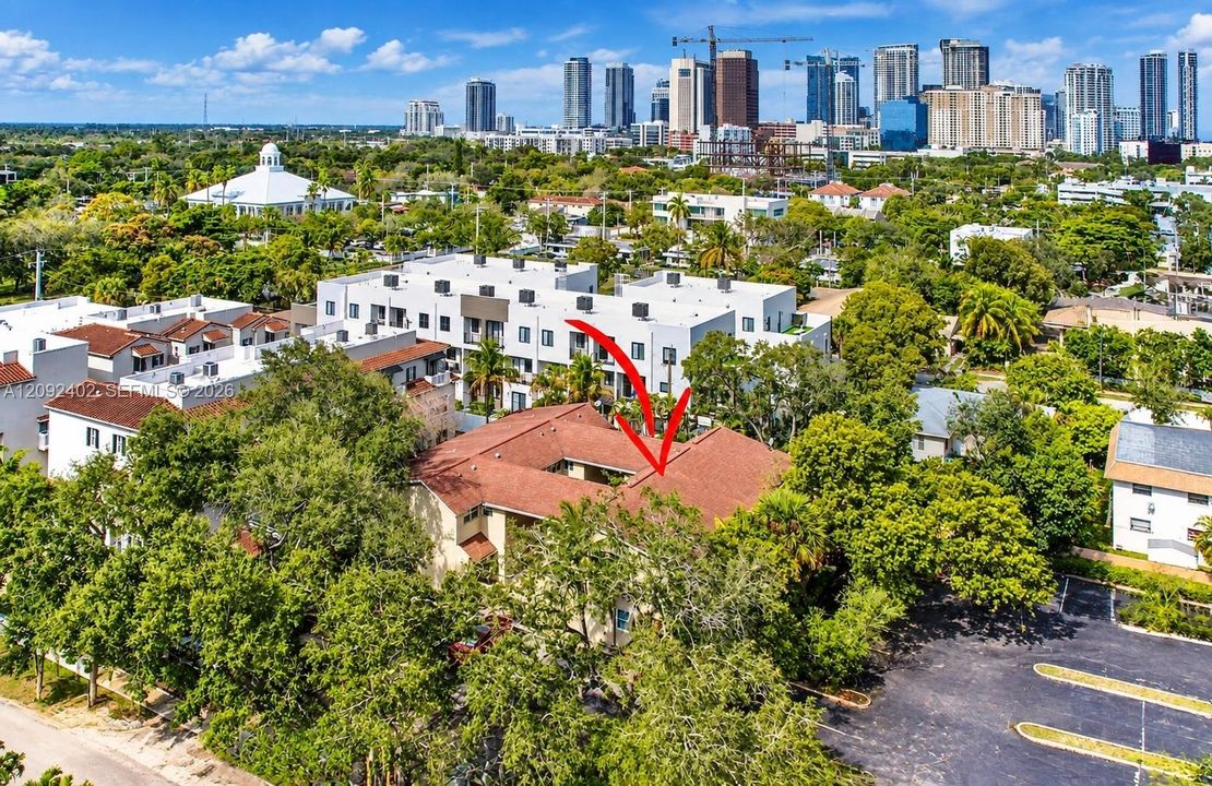 Aerial perspective of a multi-unit residential structure featuring a central courtyard, shingled roofing, and a dedicated asphalt parking area surrounded by mature trees