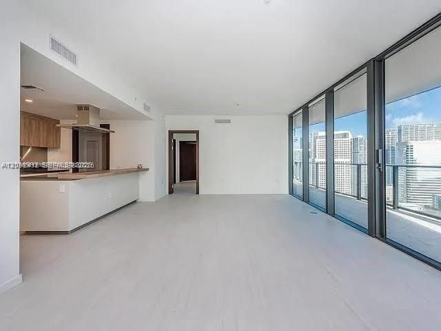 Open-concept interior featuring floor-to-ceiling glass panel walls, light-toned tile flooring, kitchen with wood-finish cabinetry, stainless steel range hood, and breakfast bar