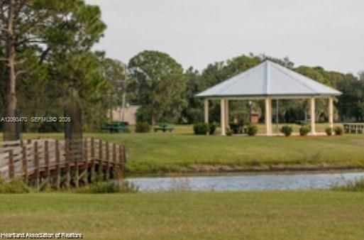Open-air gazebo with a metal roof, wooden footbridge, and expansive grass grounds surrounding a pond