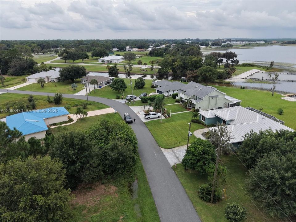 Aerial perspective of a residential neighborhood featuring homes with metal roofs, asphalt-paved streets, mature trees, open green space, and proximity to a large body of water