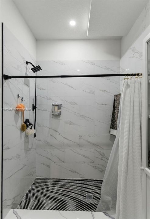 Shower enclosure featuring large-format veined wall tile, mosaic tile flooring, matte black rainfall showerhead, and wall-mounted metal soap dispenser