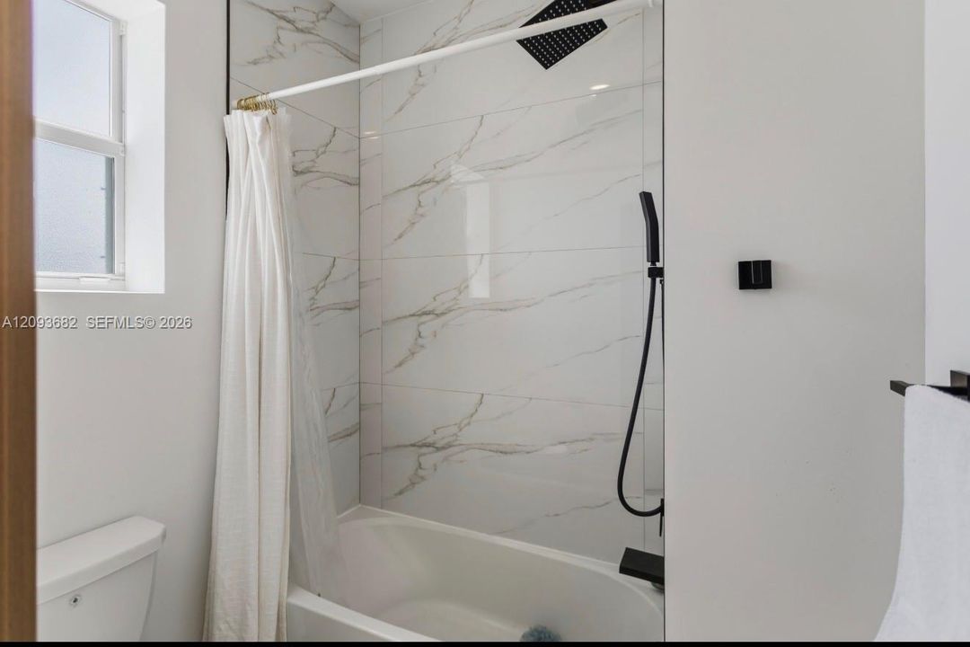 Bathtub with large-format stone-look wall tile, matte black rainfall shower head, handheld shower fixture, and matte black hardware