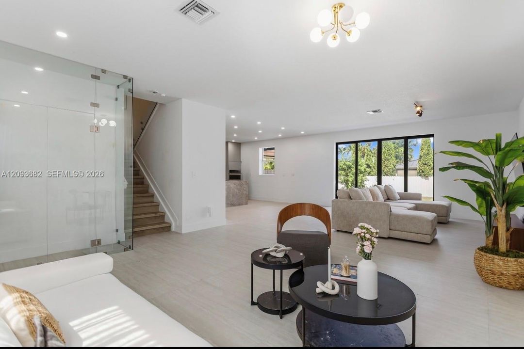 Open-concept interior featuring light-toned tile flooring, floor-to-ceiling glass wall paneling, multi-panel sliding glass doors, recessed lighting, and a modern multi-bulb ceiling fixture