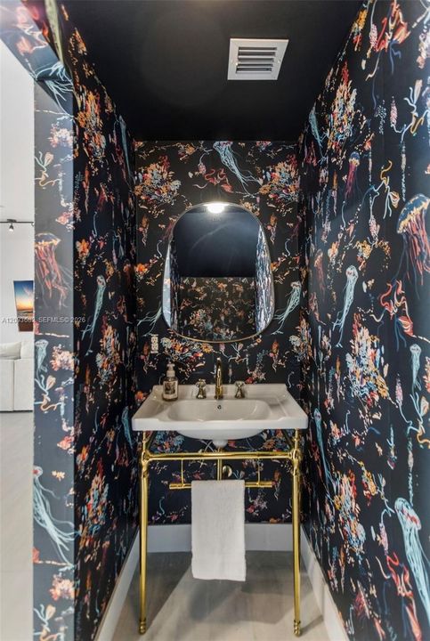 Powder room featuring bold patterned wallpaper, a wall-mounted arched mirror, and a white console sink with a metallic gold-finish frame and fixtures
