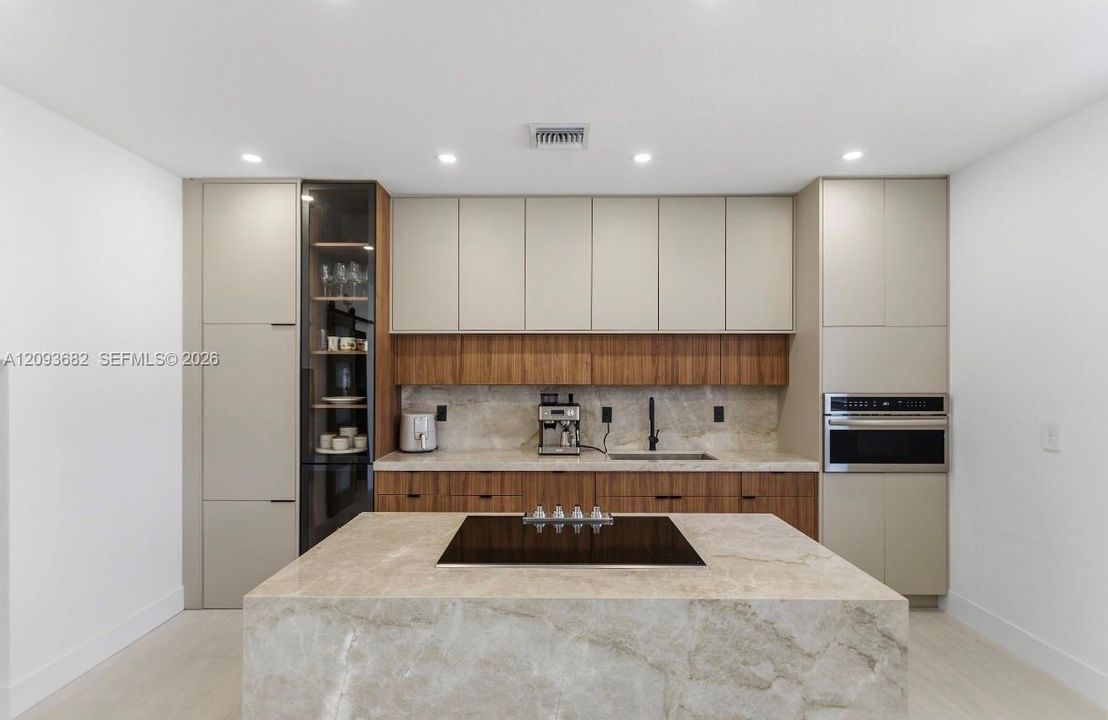 Modern kitchen featuring flat-panel cabinetry, a waterfall-edge stone island with a built-in cooktop, integrated appliances, and a glass-front display cabinet