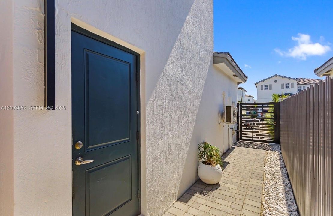 Exterior side access featuring a paved walkway, light-toned stucco wall finish, dark-toned panel door, and metal privacy fencing