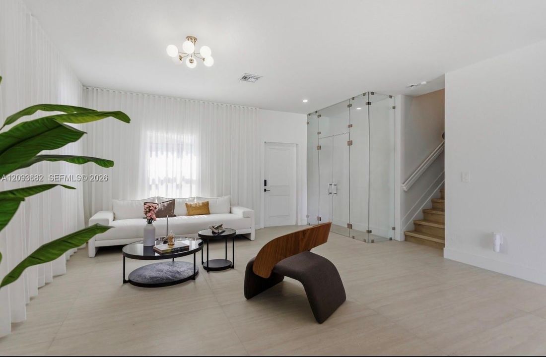 Neutral-toned tile flooring, floor-to-ceiling sheer drapery, glass-enclosed feature wall, multi-bulb modern chandelier, and wood-finish staircase
