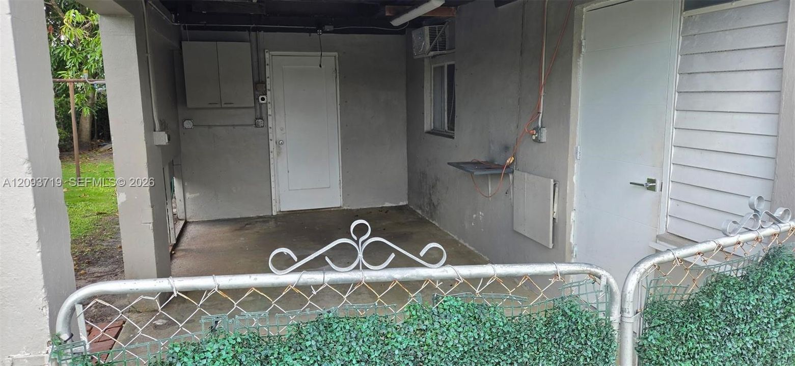 Covered carport with concrete flooring, wall-mounted storage cabinetry, two exterior entry doors, and metal chain-link perimeter fencing