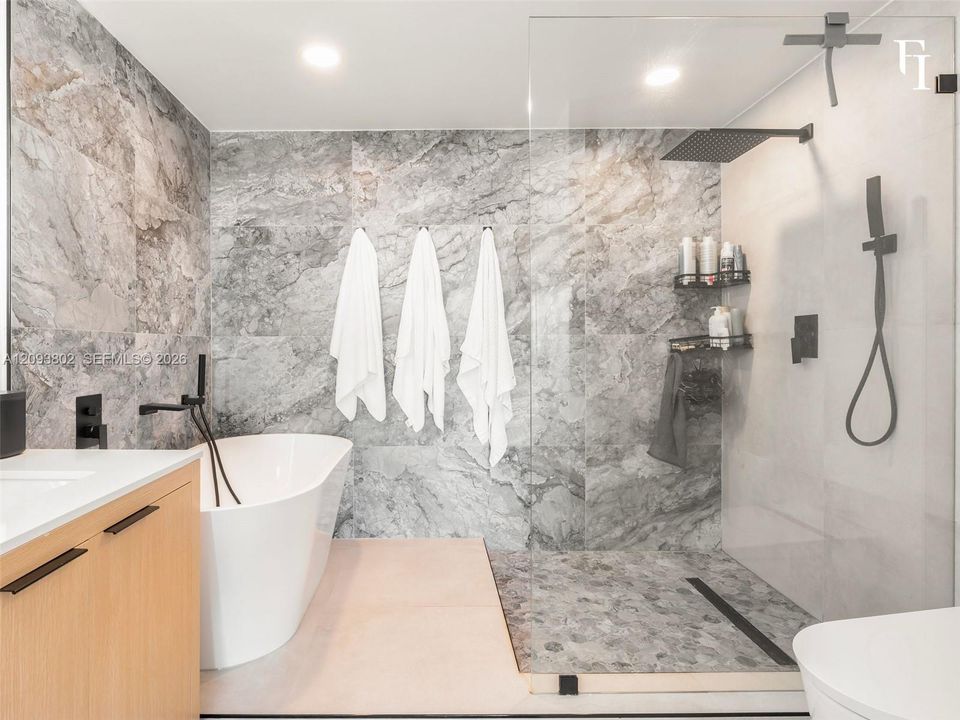Freestanding soaking tub with matte black wall-mounted faucet, walk-in shower featuring a glass enclosure and rainfall showerhead, and large-format stone-finish tile wall accents