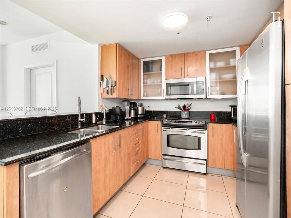 Kitchen featuring light wood cabinetry, dark stone countertops, stainless steel appliances, glass-front upper cabinet doors, and tile flooring