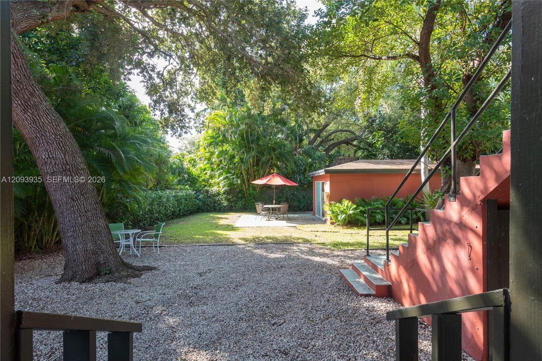 Private backyard featuring mature trees, manicured lawn, gravel surface, and exterior staircase with black metal railings