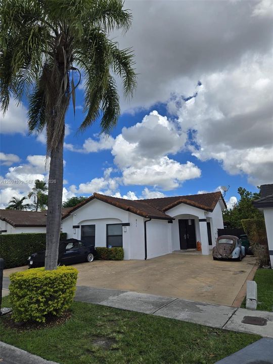 Single-story exterior featuring white stucco finish, a tile roof, a paved driveway, and established palm and shrub landscaping