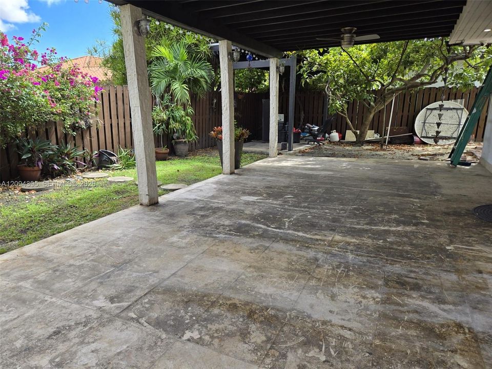 Covered patio with a beamed ceiling, stone-finish flooring, and supporting columns, complemented by a fenced backyard featuring mature trees and established landscaping