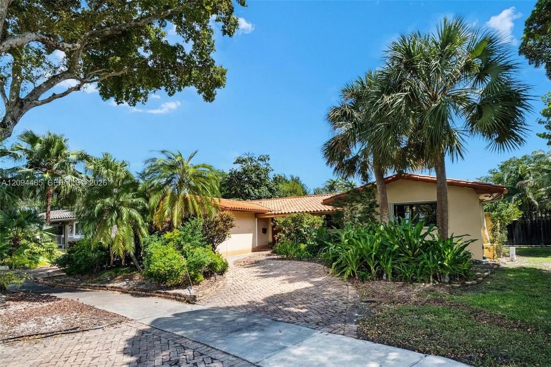 Single-story residence featuring a terracotta-tiled roof, stucco exterior, and a herringbone-patterned brick paver driveway, surrounded by established landscaping and mature palm trees