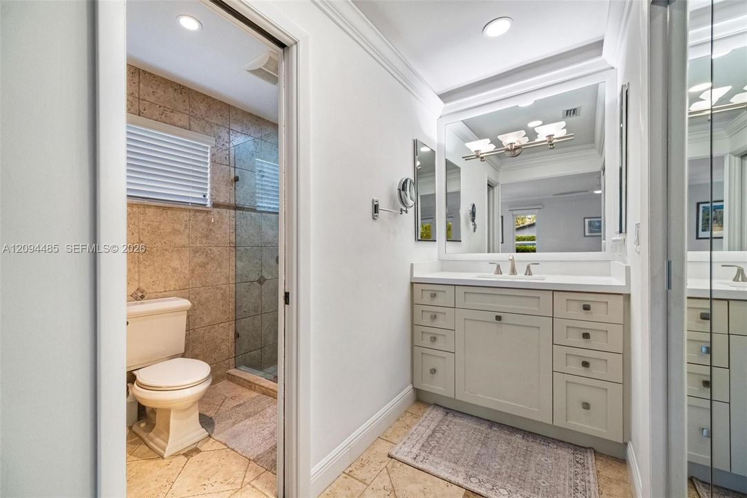 Vanity featuring a white countertop with an integrated basin, shaker-style cabinetry with metallic hardware, large-format wall mirror, and natural stone tile flooring