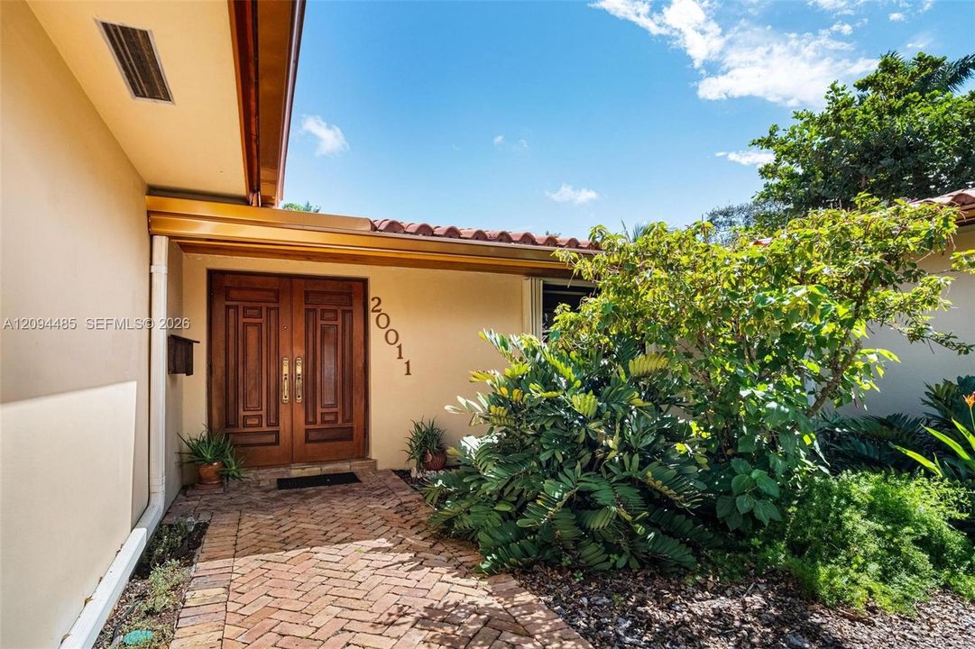 Stucco exterior entry featuring a herringbone-patterned brick walkway, double wood-paneled doors with metallic hardware, and a terracotta tile roof edge