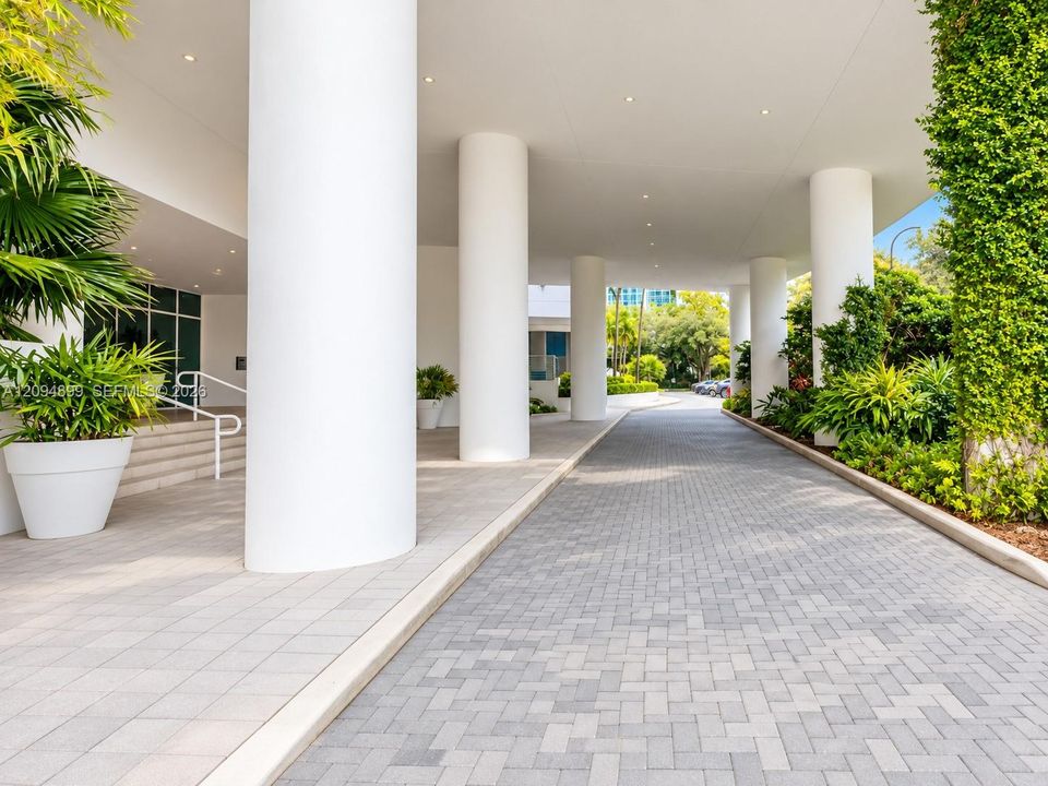 Covered motor court featuring cylindrical support columns, herringbone-patterned paver driveway, recessed ceiling lighting, and manicured perimeter landscaping