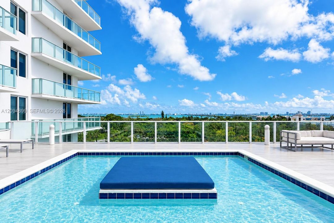 Rooftop swimming pool featuring a central raised platform, tiled perimeter, light-toned paved decking, and glass panel railings