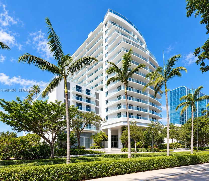 Multi-story residential building featuring a curved architectural design, stacked glass-railed balconies, exterior stucco facade, and manicured landscaped grounds
