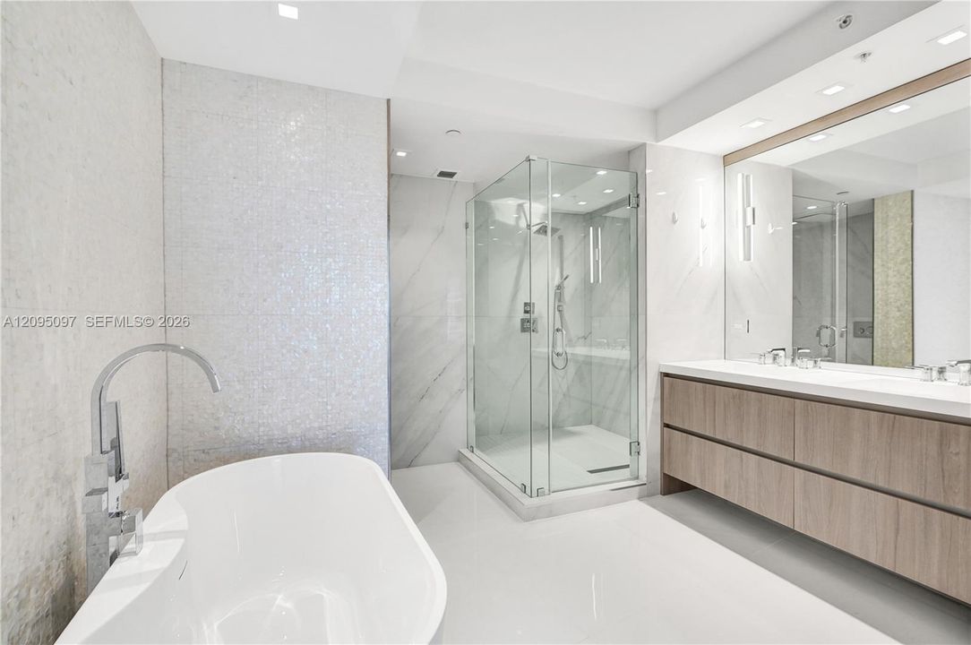 Freestanding soaking tub with floor-mounted faucet, glass-enclosed shower, wall-to-wall vanity with double sinks, and recessed lighting