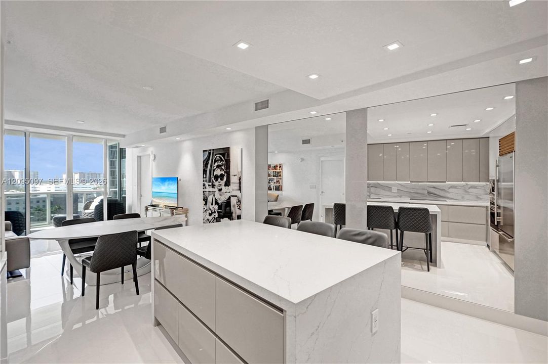 Open-concept interior featuring high-gloss cabinetry, a central island with a solid surface countertop, stone-finish backsplash, recessed ceiling lighting, and floor-to-ceiling windows