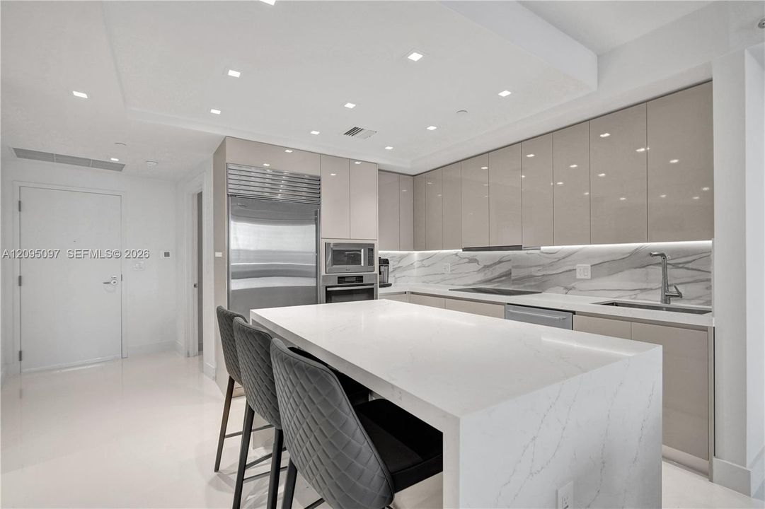 Contemporary kitchen featuring high-gloss cabinetry, a waterfall-edge quartz island, under-cabinet lighting, integrated stainless steel appliances, and stone-finish backsplash