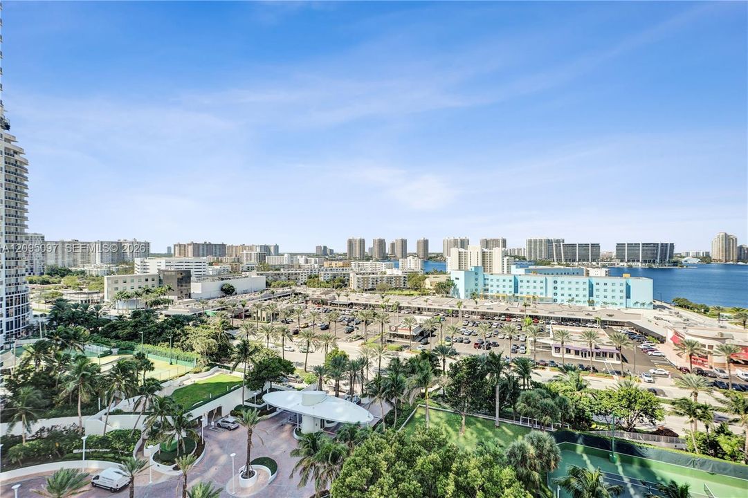 Elevated exterior perspective featuring a circular porte-cochère, paved motor court, mature landscaping, and tennis courts