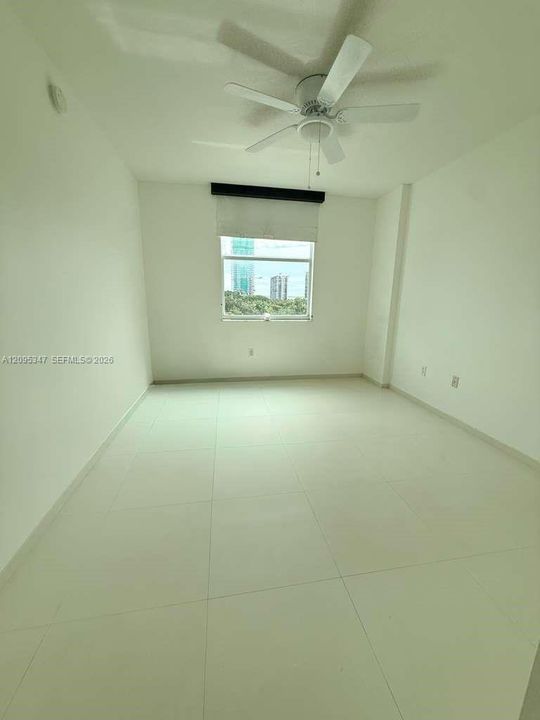 Neutral-toned room featuring large-format tile flooring, a white ceiling fan, and a single window with a roller shade