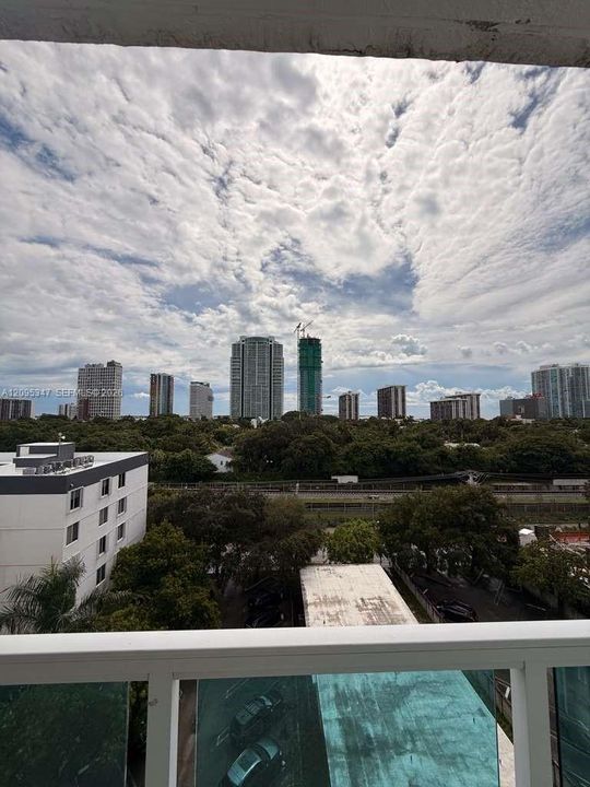 Balcony featuring a glass and white-finished metal railing overlooking a cityscape skyline, mature trees, and ground-level parking