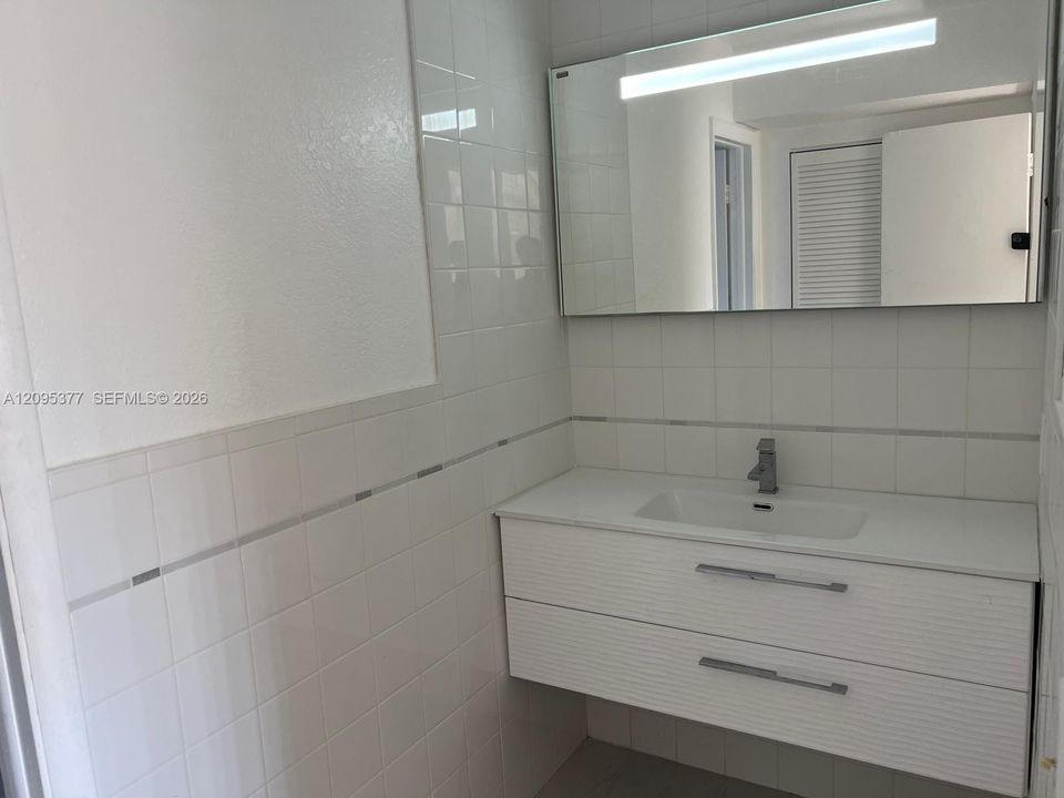 Floating vanity with a white countertop and integrated sink, complemented by a large wall-mounted rectangular mirror featuring an integrated light fixture and white tiled walls