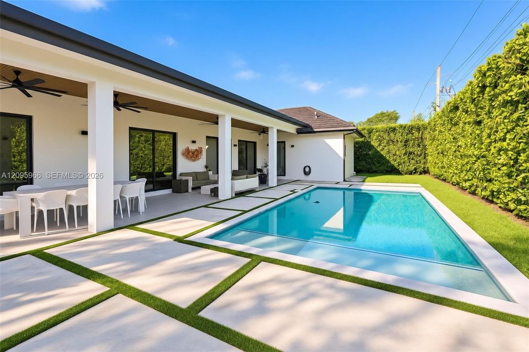 Covered outdoor patio featuring recessed ceiling fans, rectangular concrete pavers with grass inlay, and a rectangular swimming pool bordered by a tall privacy hedge