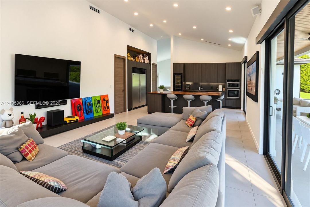 Open-concept interior featuring vaulted ceilings with recessed lighting, large-format tile flooring, kitchen with dark-finish cabinetry and integrated appliances, and a wall of glass sliding doors