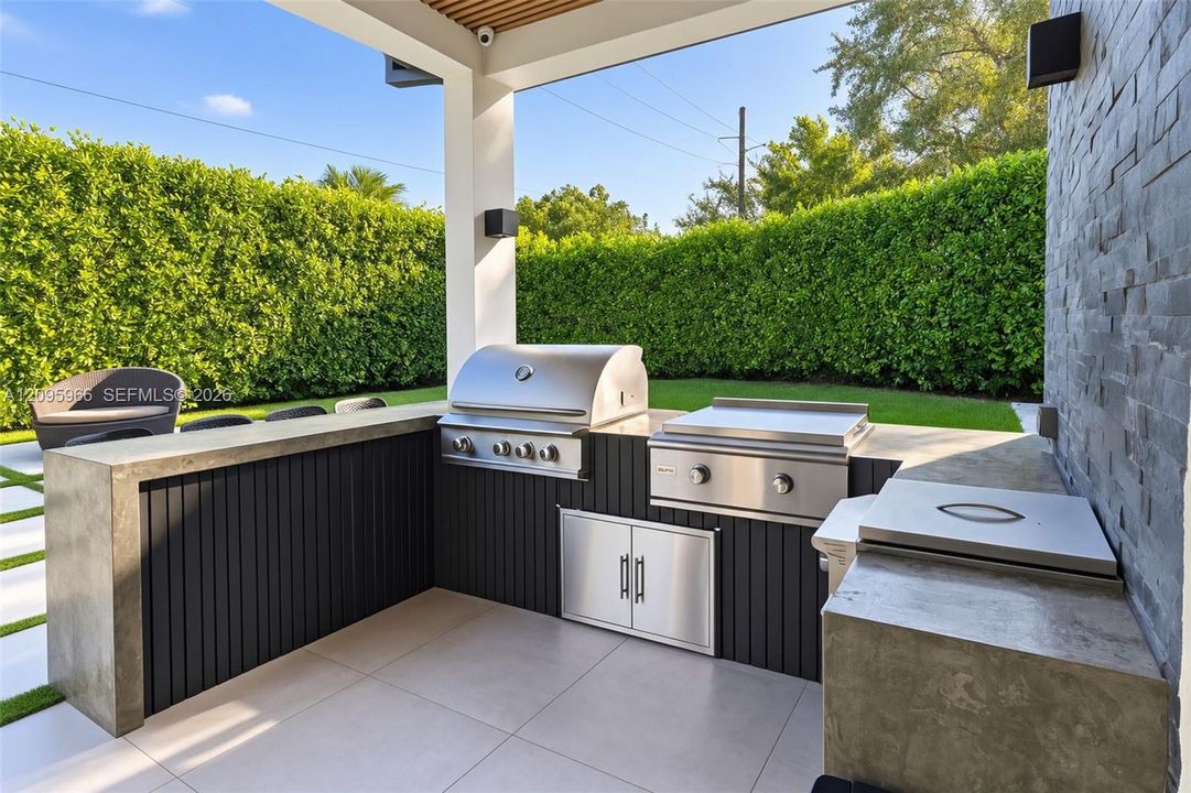 Covered outdoor kitchen featuring a built-in stainless steel grill, a flat-top griddle, concrete countertops, vertical slat cabinetry, and a stacked stone accent wall