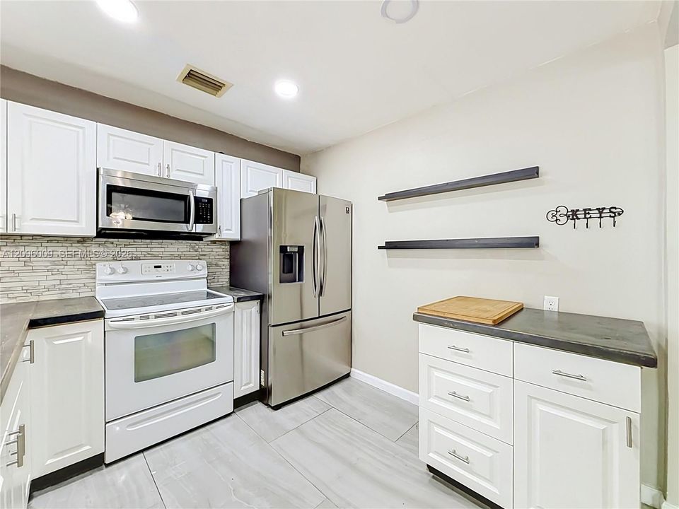 Kitchen featuring white cabinetry, stainless steel appliances, linear tile backsplash, dark solid-surface countertops, and light-toned tile flooring