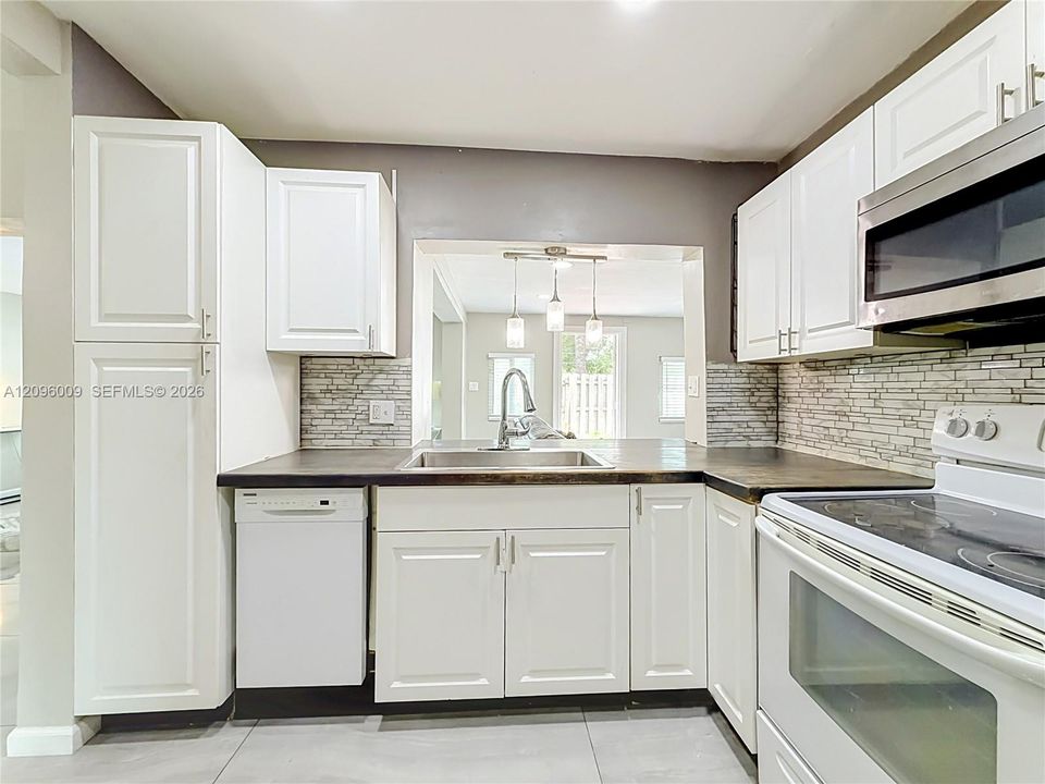 Kitchen featuring white cabinetry, dark countertops, a stacked-stone tile backsplash, stainless steel sink with pull-down faucet, and white appliances