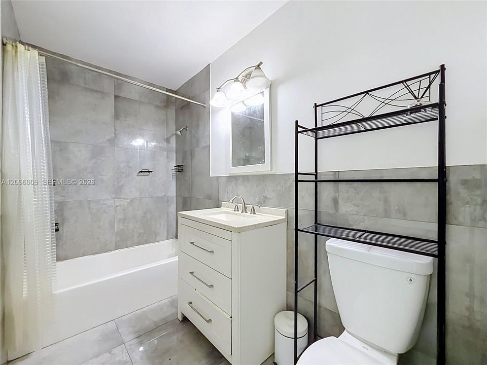 Bathroom featuring large-format grey tile surround, white vanity cabinet with stone-finish countertop, undermount sink, and three-light wall fixture
