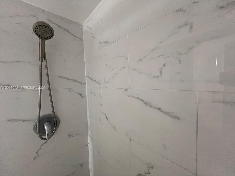Shower surround featuring large-format, light-toned stone-finish tile with integrated veining, complemented by a metallic handheld showerhead and circular wall-mounted control valve