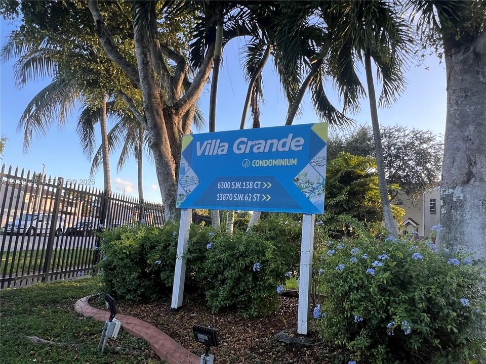 Exterior signage for Villa Grande Condominium featuring mature palm trees, perimeter metal fencing, and landscaped shrubbery