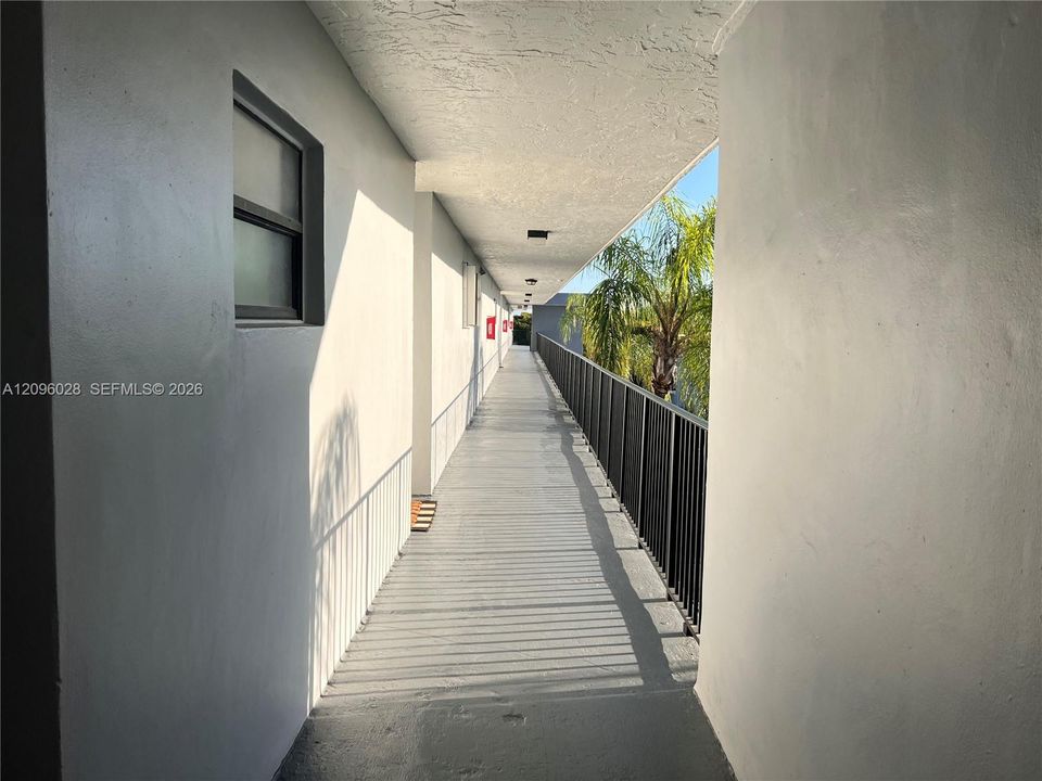 Exterior walkway featuring a painted concrete floor, solid masonry walls, a metal railing, and surface-mounted ceiling light fixtures