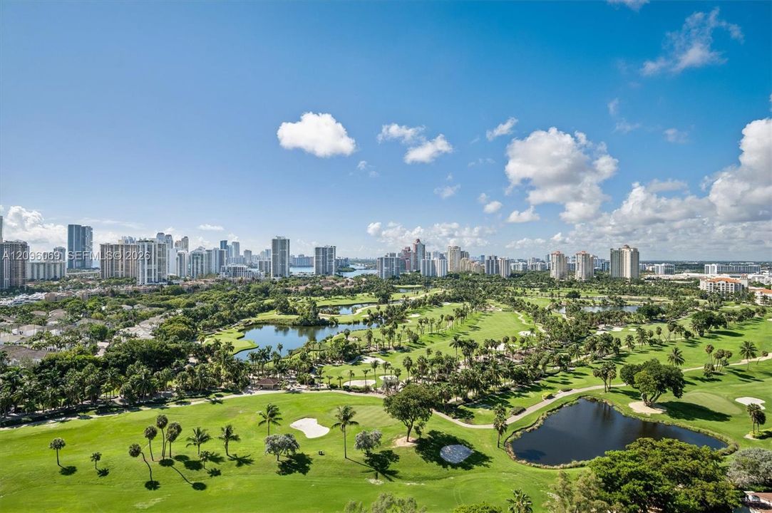 Panoramic aerial perspective featuring expansive golf course fairways, multiple water features, mature tropical vegetation, and a metropolitan skyline backdrop