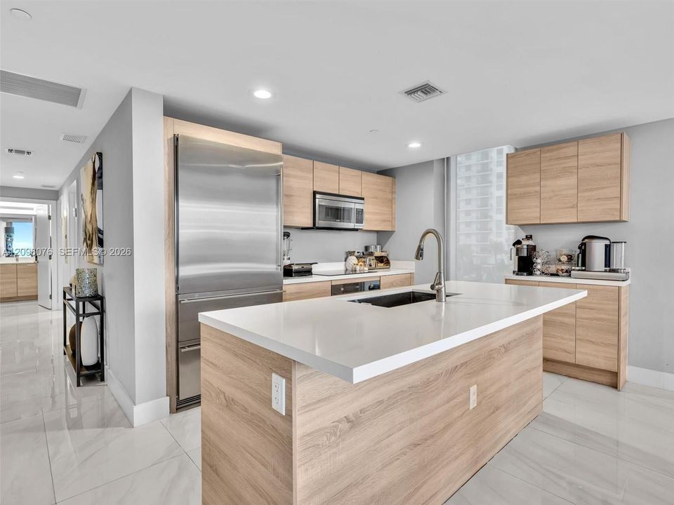 Kitchen featuring a central island with a quartz countertop and under-mount sink, light-tone wood-finish cabinetry, stainless steel refrigerator, and large-format tile flooring