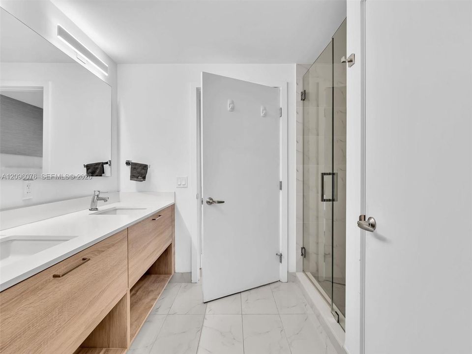 Bathroom featuring a dual-sink vanity with light wood-finish cabinetry, solid surface countertops, a full-width mirror, and a walk-in shower with a glass enclosure and tile flooring