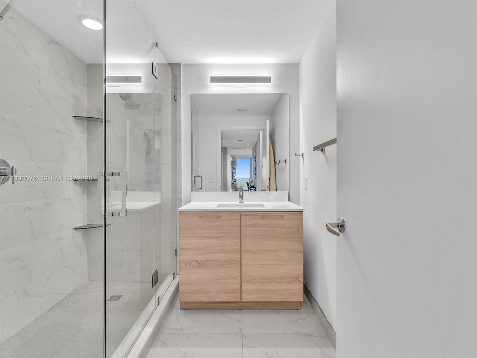 Walk-in shower with frameless glass enclosure, wood-finish cabinetry, white countertop with integrated sink, and large-format tile flooring