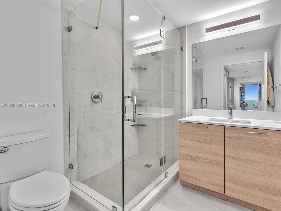 Walk-in glass-enclosed shower with stone-finish tile surround, light wood-finish cabinetry, white countertop with integrated sink, and large vanity mirror with wall-mounted lighting