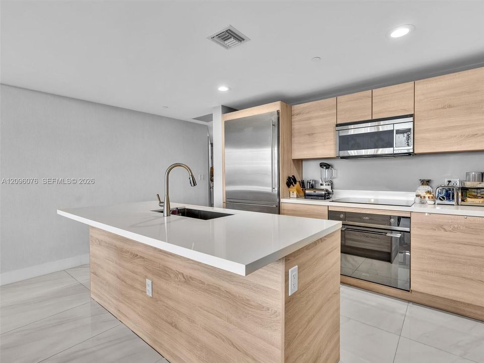 Kitchen featuring flat-panel wood-finish cabinetry, white composite countertops, a center island with an undermount sink, stainless steel appliances, and large-format tile flooring